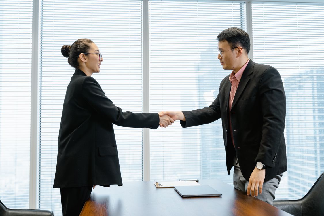 A man and woman shaking hands across a desk in a professional meeting